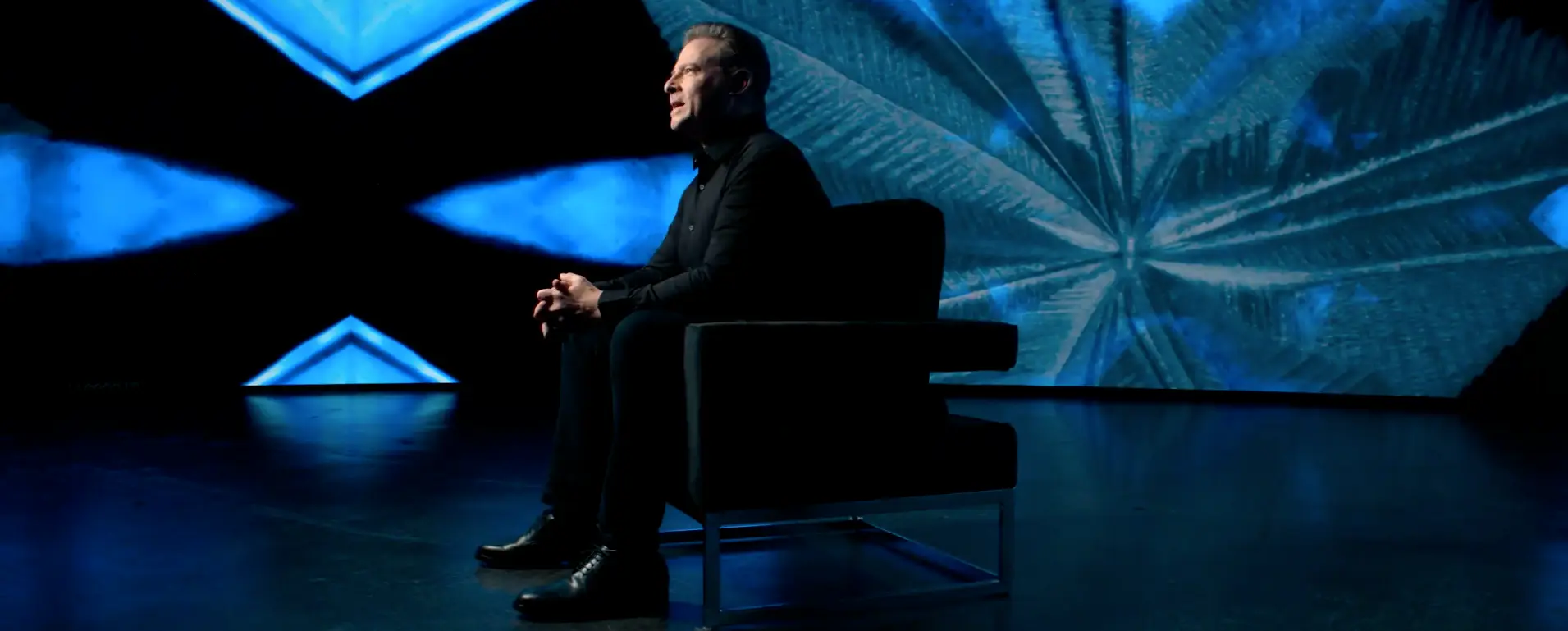 David Schmidt seated in a studio interview setting with abstract geometric visuals in the background